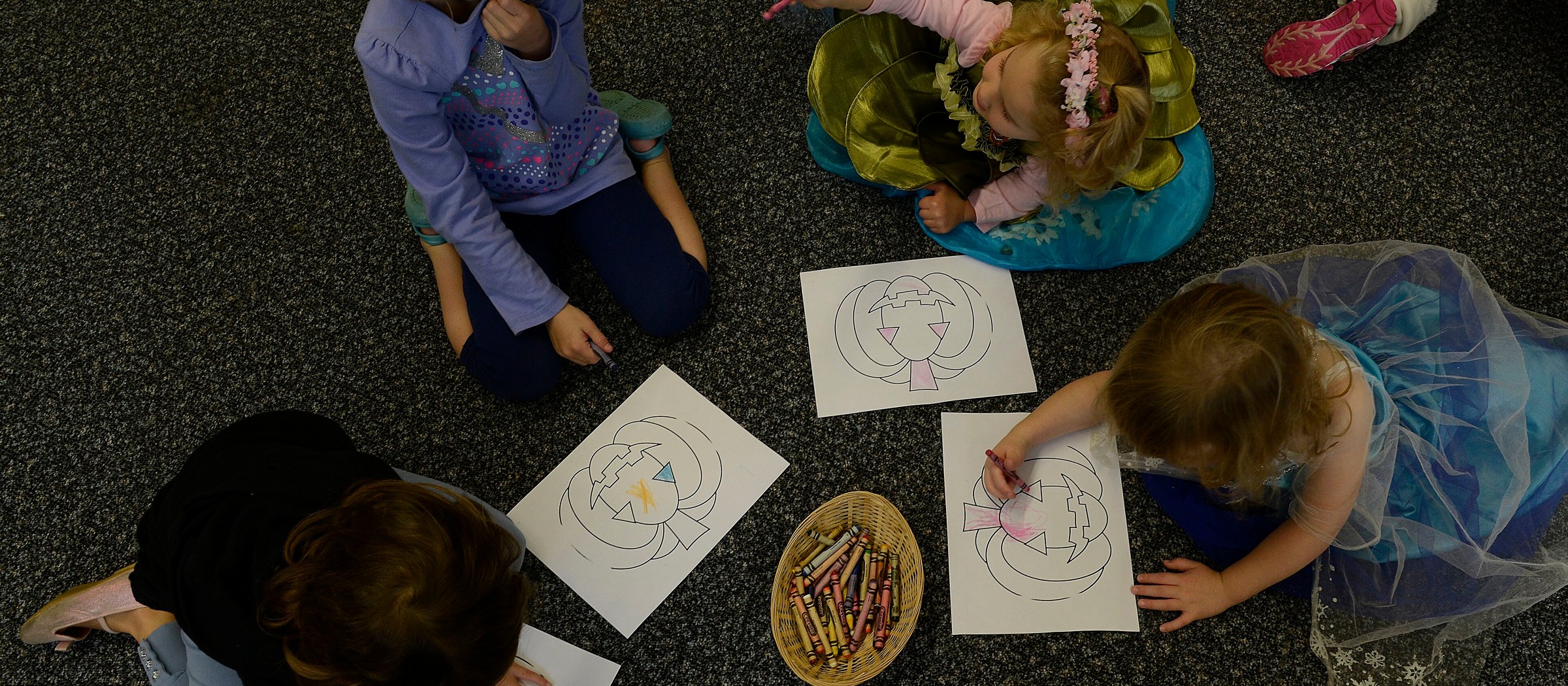 Cuatro niños pequeños disfrazados, sentados en la alfombra de una biblioteca, colorean dibujos impresos con una canasta de crayones entre ellos