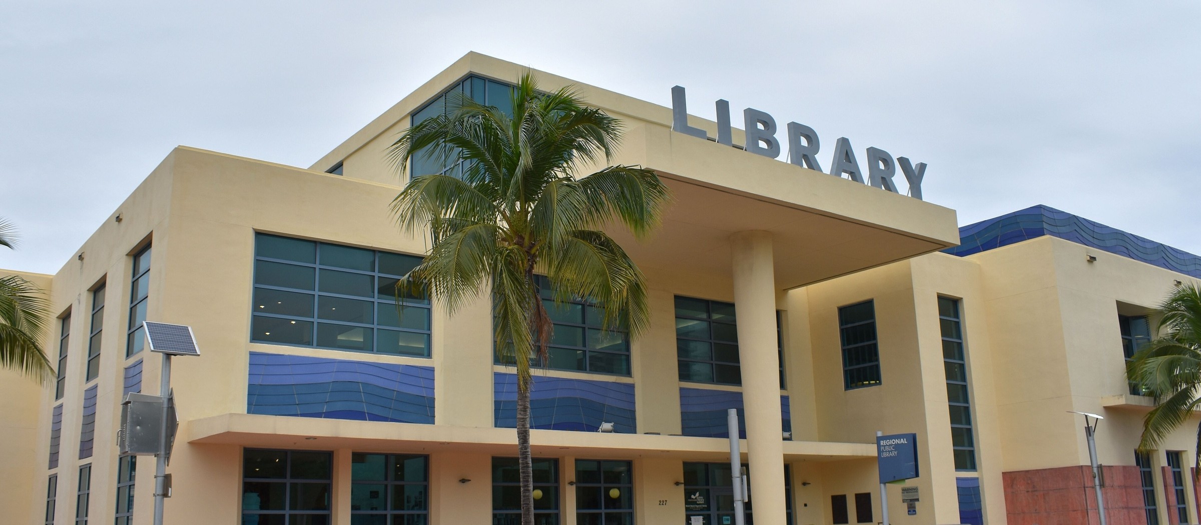 La Miami Beach Regional Library, un edificio color crema con franjas azules en las ventanas y la palabra 'LIBRARY' en letras grandes sobre la entrada, detrás de una palmera