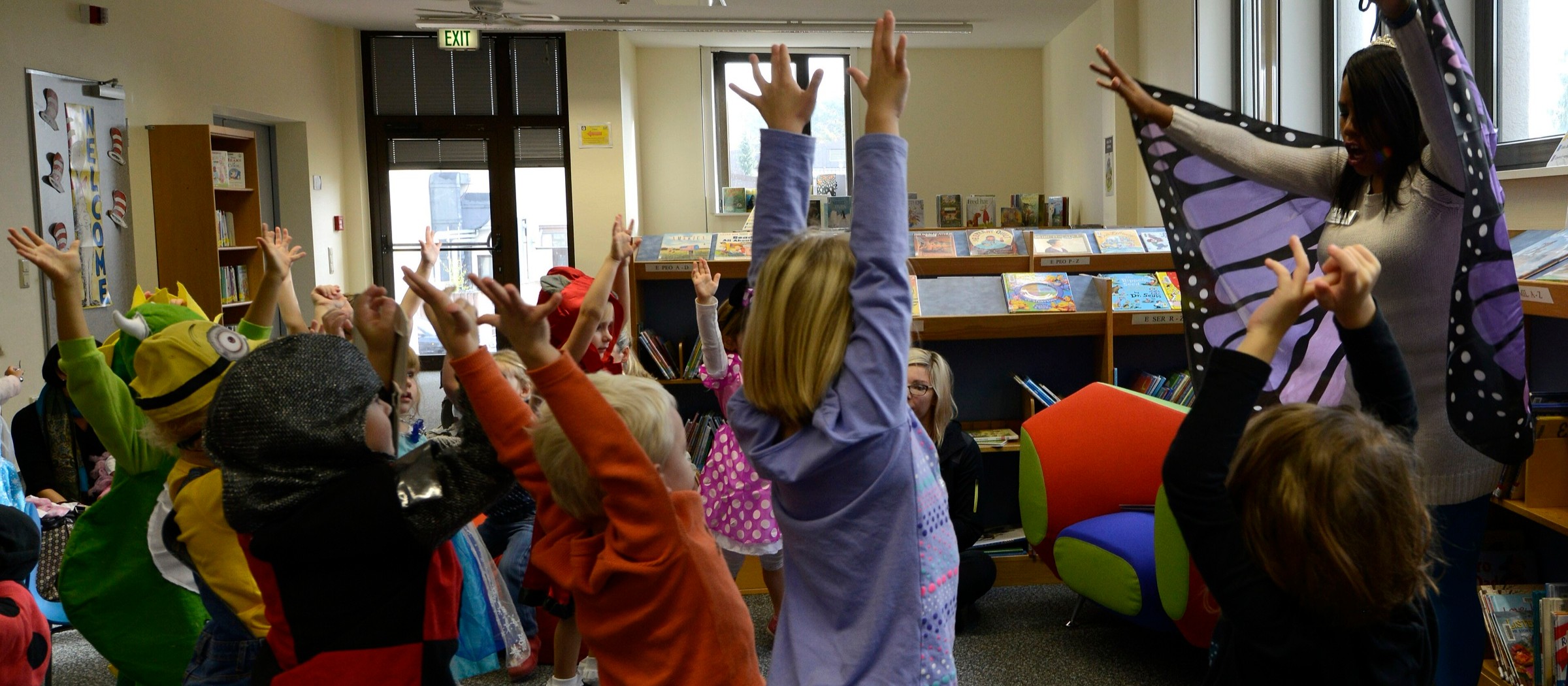 Niños disfrazados de Halloween levantan los brazos en una canción que dirige una bibliotecaria con alas de mariposa