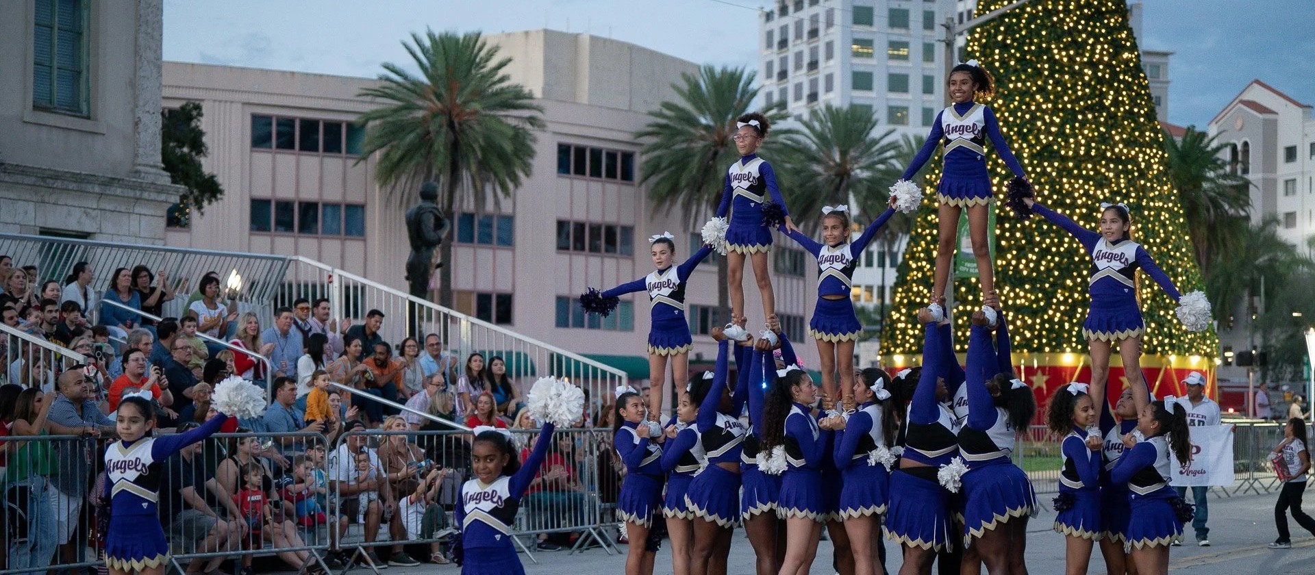 Jóvenes porristas de azul y blanco actúan frente a un árbol de Navidad iluminado mientras el público mira el desfile