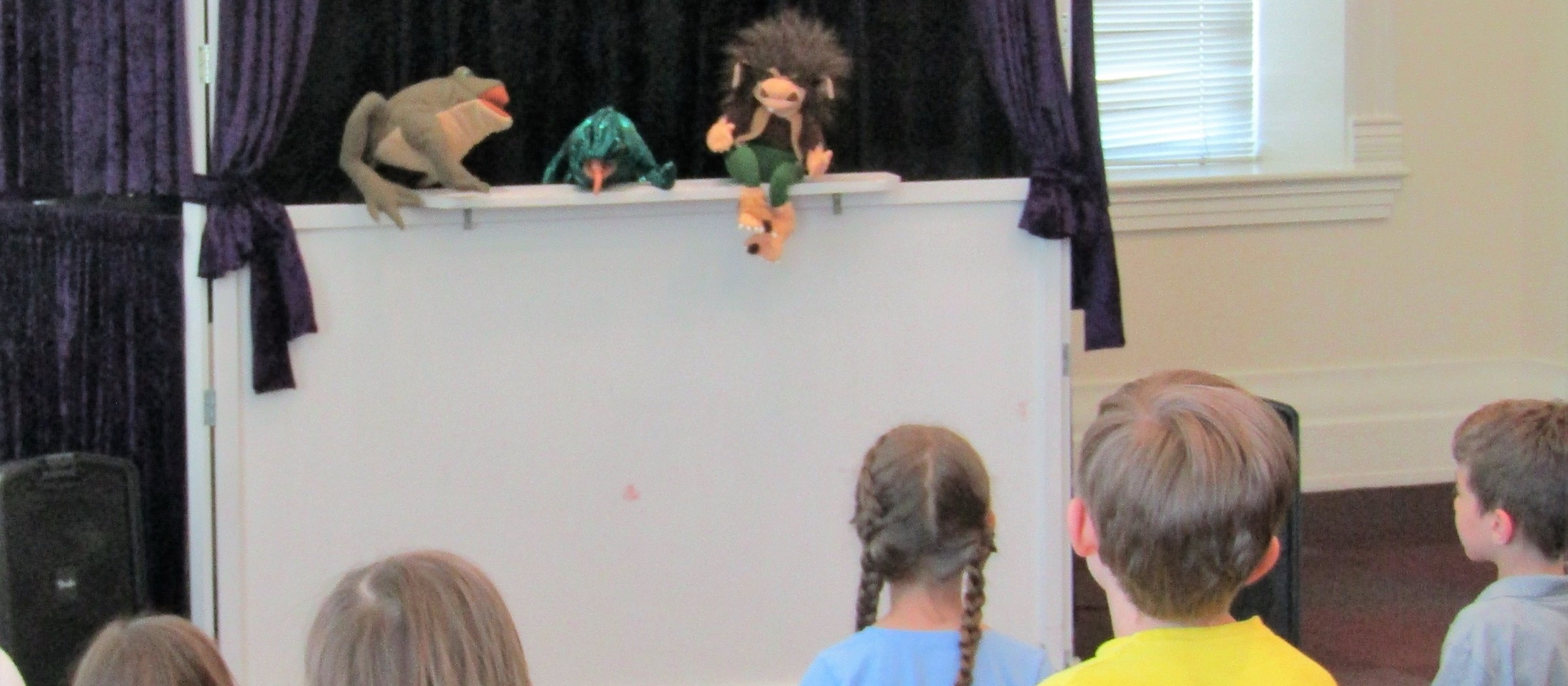 Children seen from behind watching a puppet show on a small stage with purple curtains