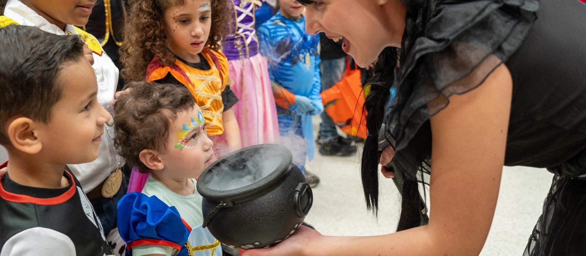 Children in Halloween costumes peering into a smoking cauldron held by a woman dressed as a witch