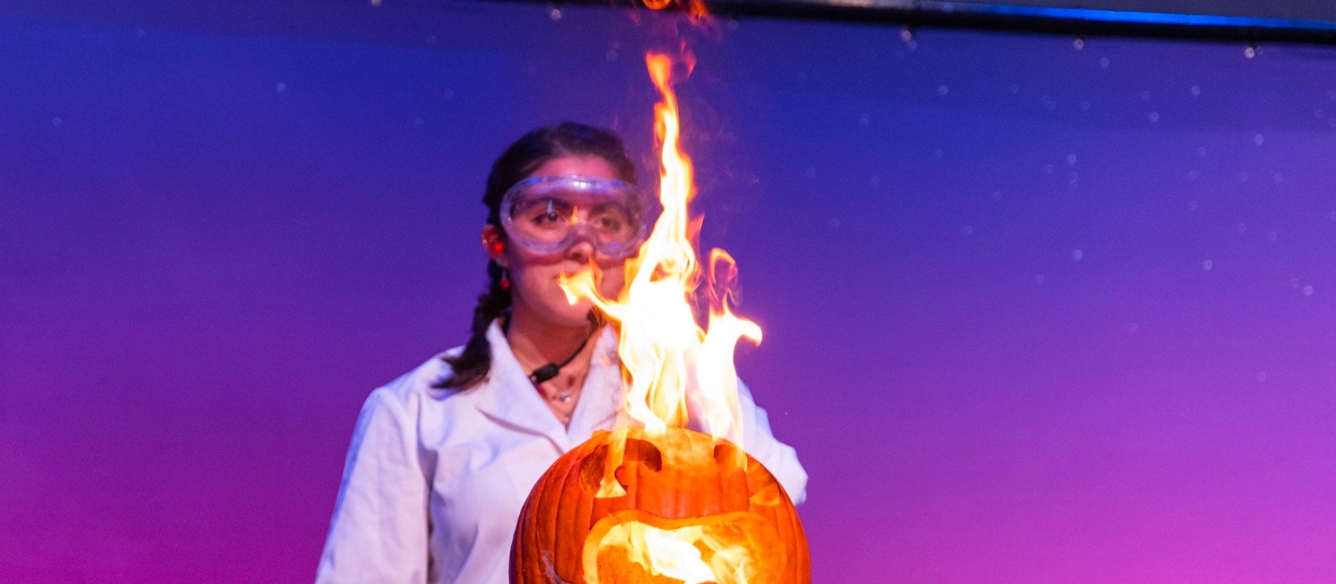 Una científica con gafas de seguridad y bata de laboratorio detrás de una calabaza de Halloween que estalla en llamas durante un espectáculo de química