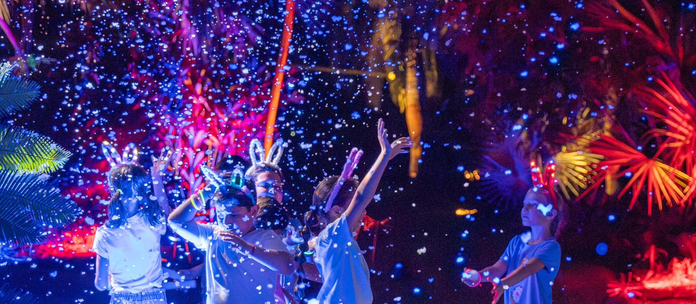 Children in light-up bunny ears dance under falling bubbles and colored lights at NightGarden