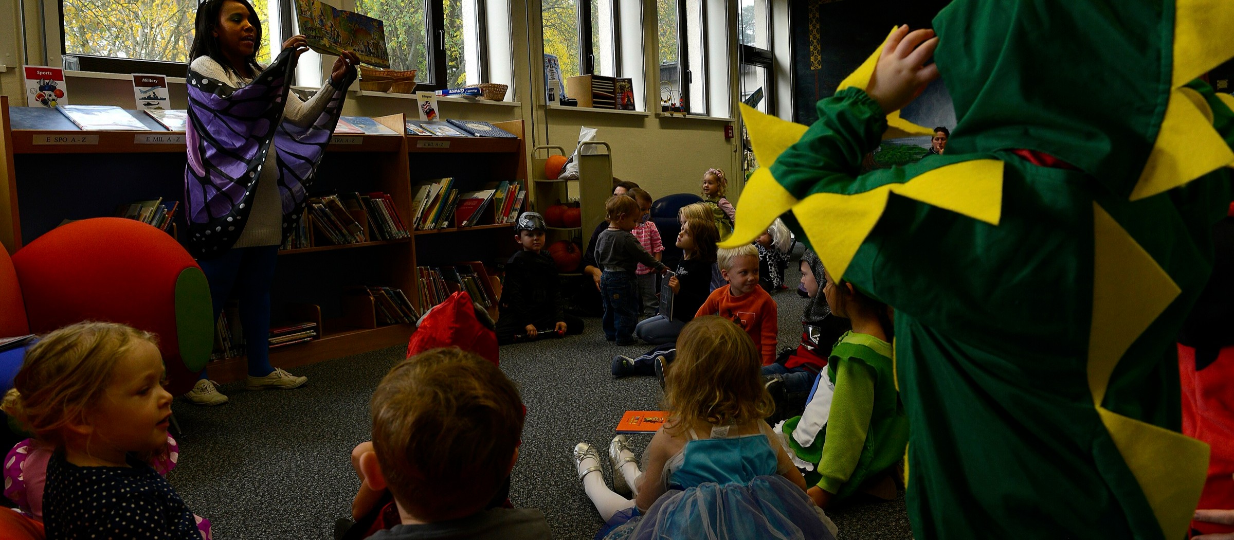 Una bibliotecaria con alas de mariposa lee un libro ilustrado a niños pequeños disfrazados de Halloween sentados en el suelo de una biblioteca