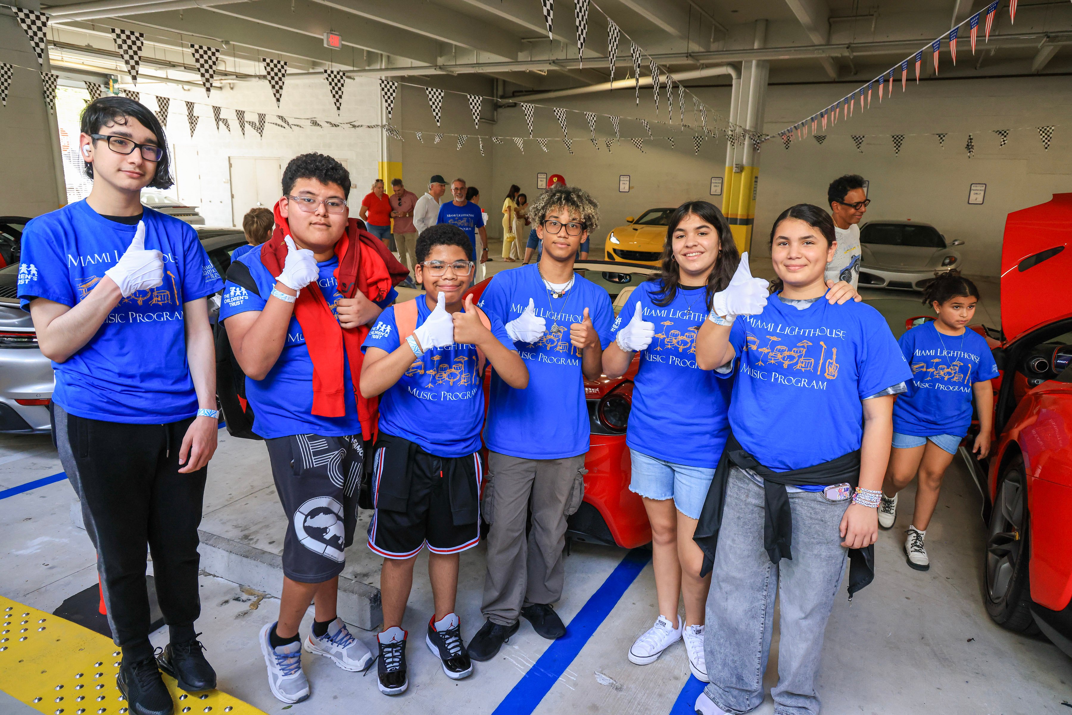 Music Program students in blue T-shirts and white gloves giving a thumbs-up among sports cars under checkered pennants