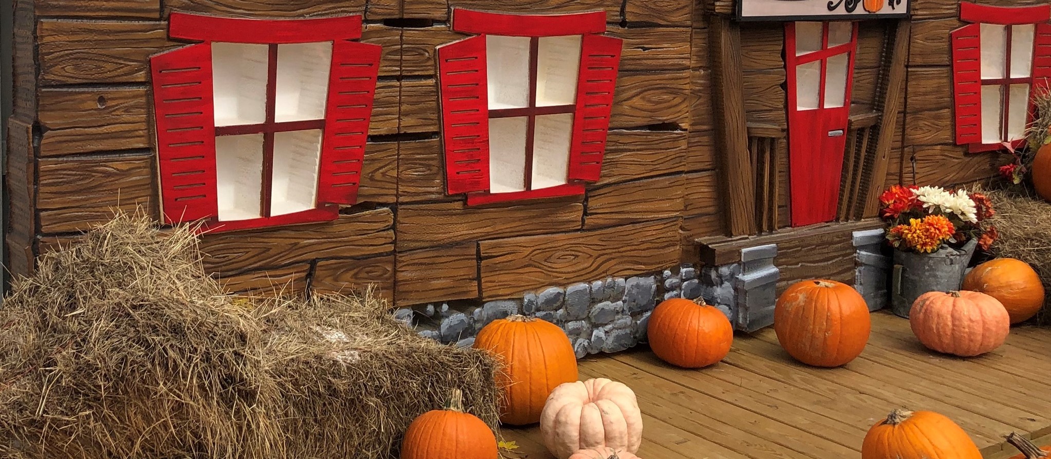 A storybook-style wooden cottage with an orange tile roof, red shutters and a 'Pumpkin Patch' sign, with pumpkins and hay bales in front