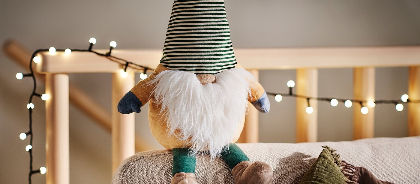 A plush gnome in a tall green-striped hat sitting on a sofa arm, string lights behind