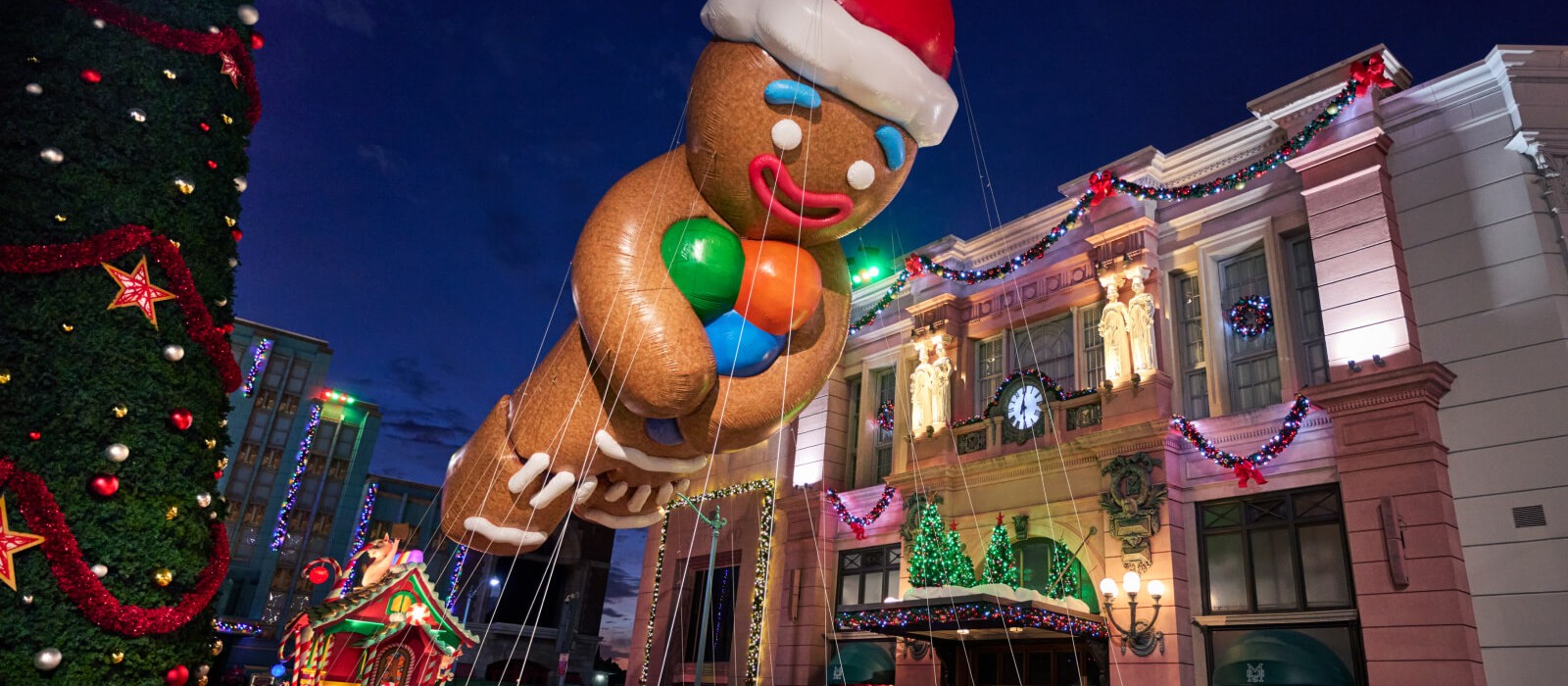A giant Gingerbread Man balloon in a Santa hat floating past decorated buildings