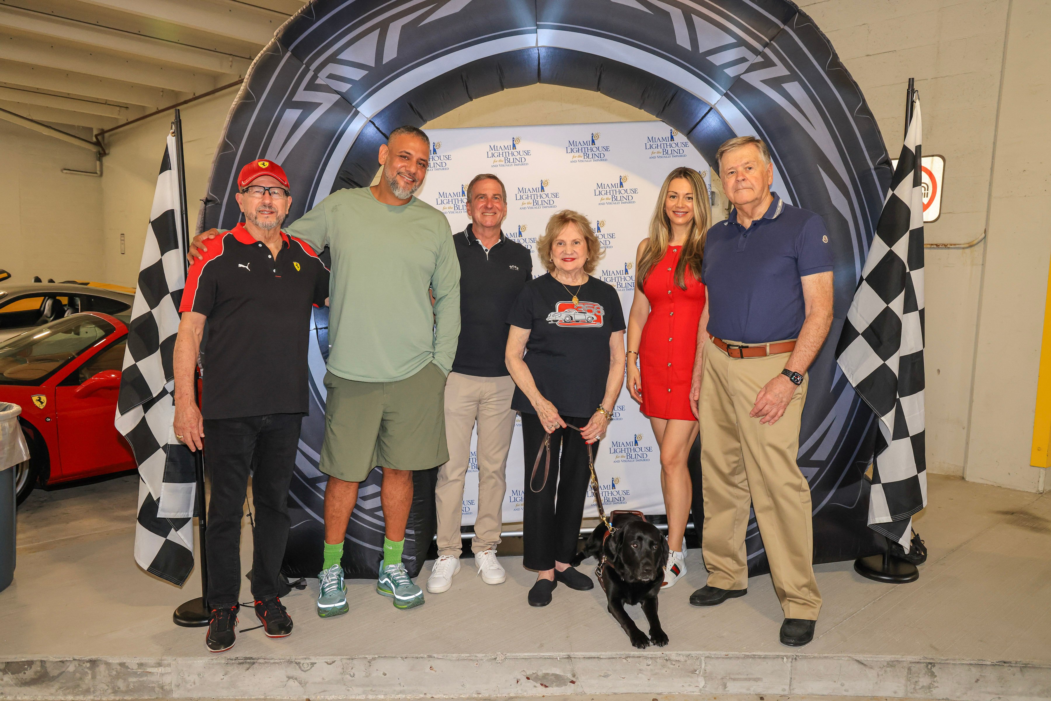 Six board members in front of an inflatable racing-tire arch between two checkered flags; Virginia Jacko holds her black guide dog on a leash