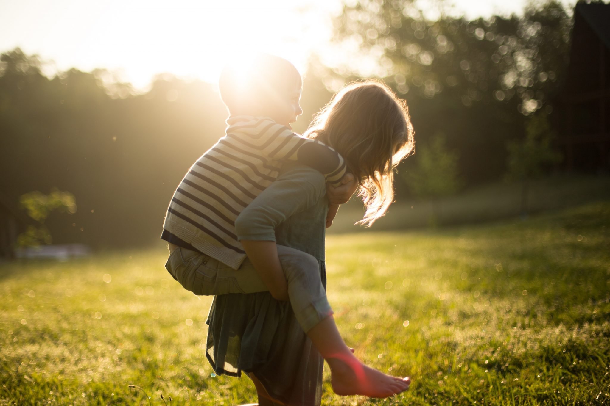A child carrying a younger child piggyback across a sunlit lawn