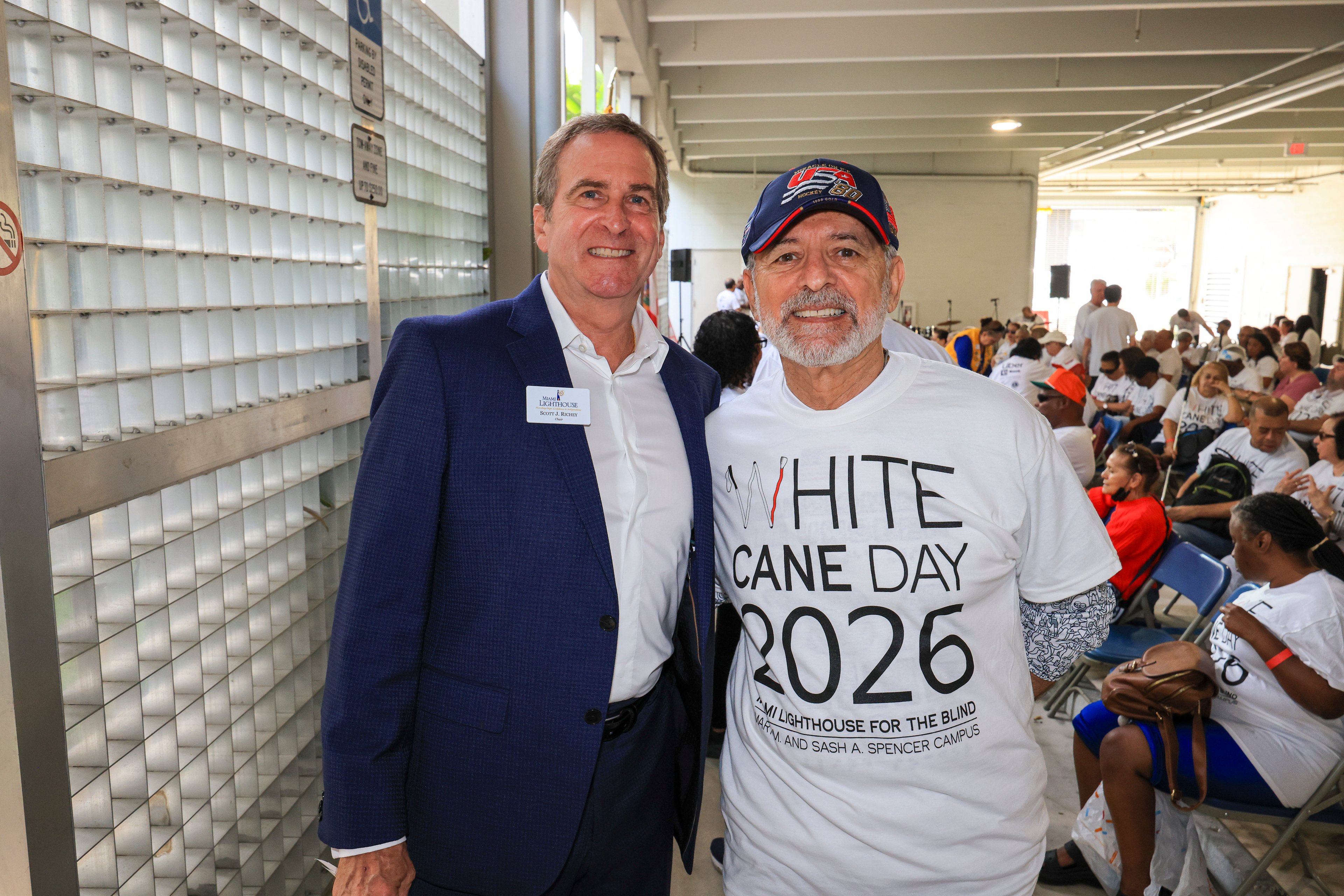 Scott J. Richey con saco azul marino y Thomas Abraham con camiseta y gorra del White Cane Day 2026, con invitados sentados detrás