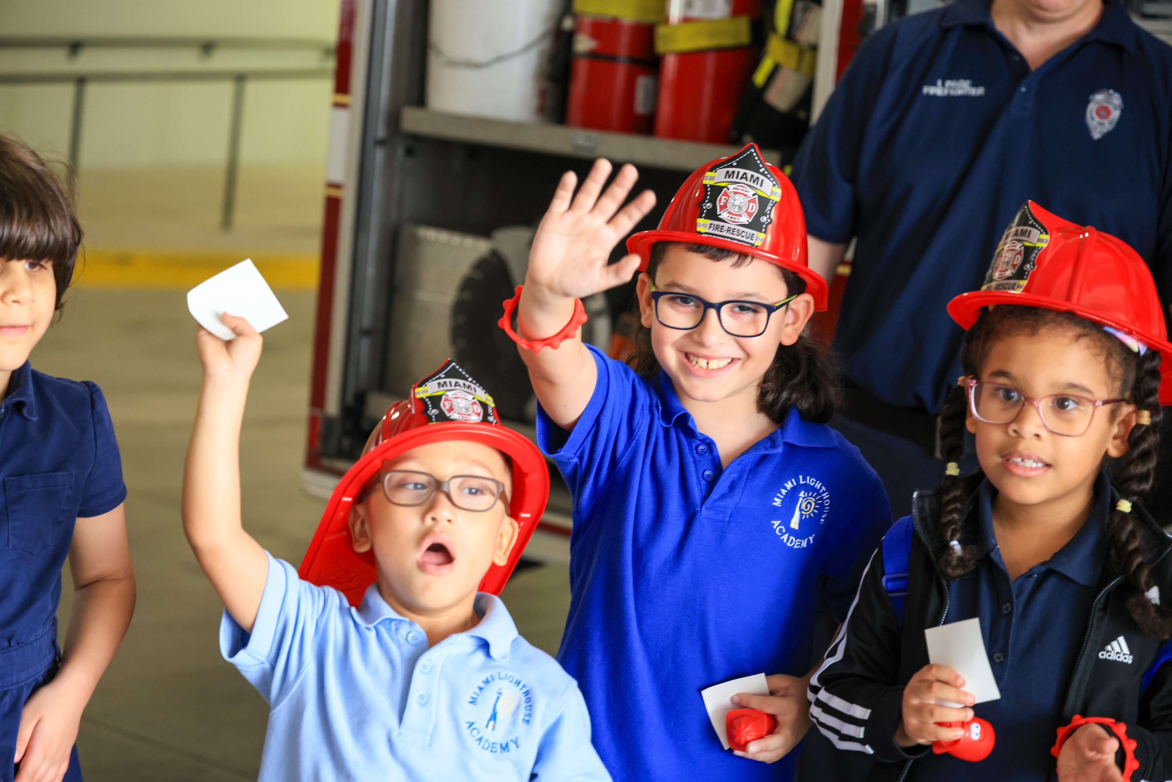 Estudiantes de Miami Lighthouse Academy con cascos de bombero rojos de juguete saludan junto a un camión de bomberos