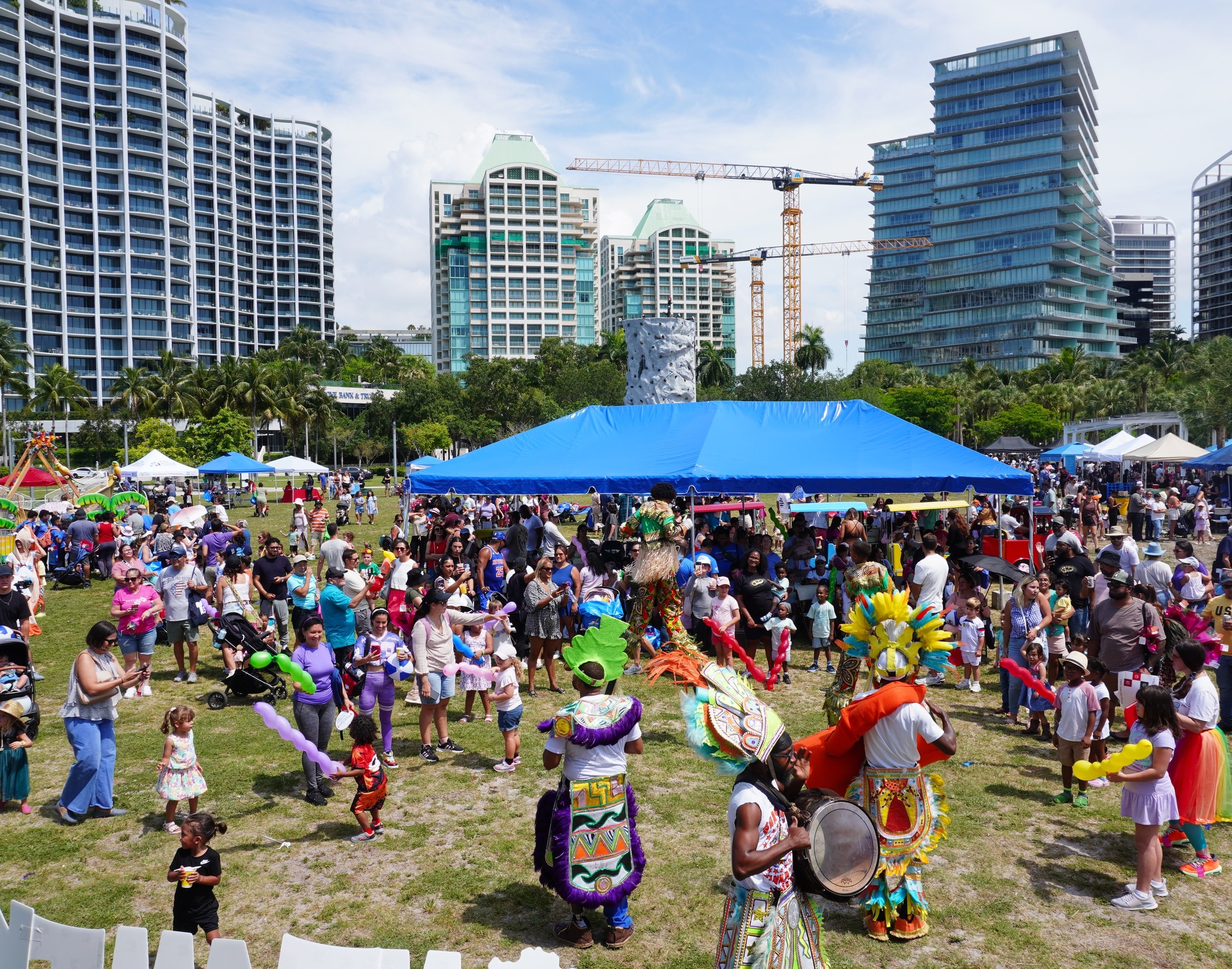 Families and costumed performers on a park lawn at Miami Kids Magazine's Family Fest, with Miami high-rises behind