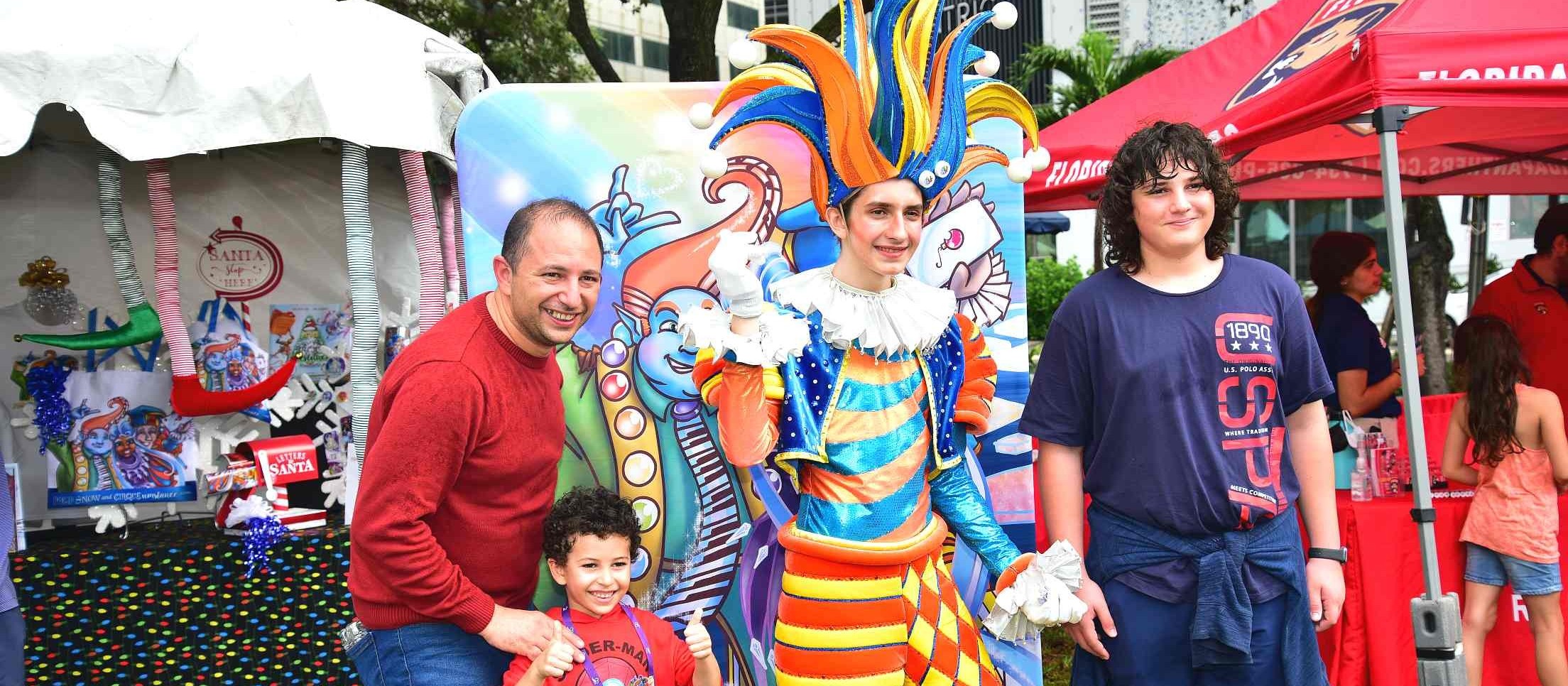 Un padre, un niño, un adolescente y un artista disfrazado de bufón de colores posan frente a un mural pintado en el Family Fun Day