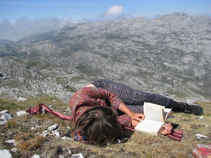 A person resting beside an open book on a grassy mountain slope.
