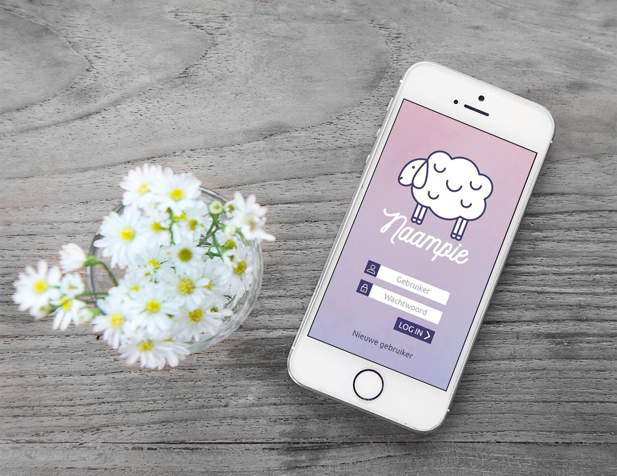 A white iPhone lying on grey weathered wood beside a glass of white daisies, showing the Naampie login screen: a sleeping sheep in outline, the name in white script, fields for username and password, a log in button and a link for new users.