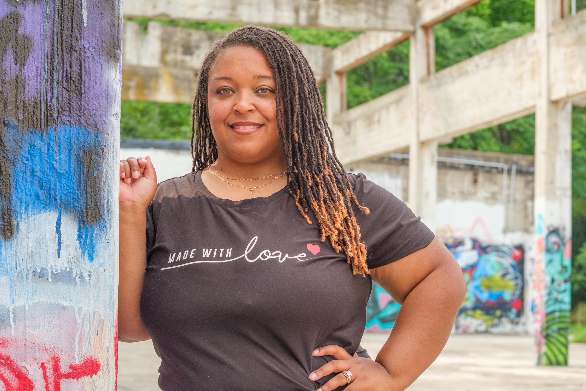 A woman in a Made With Love tee leaning against a paint-streaked column, the open concrete structure and green trees behind her.