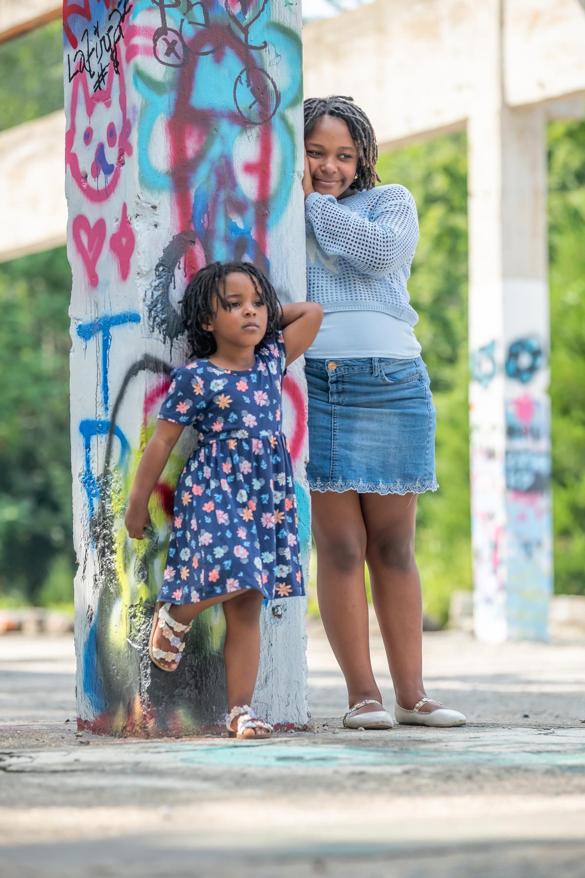 Charlie and Cori either side of a graffiti-covered column, Cori resting her cheek against it, green trees behind them.