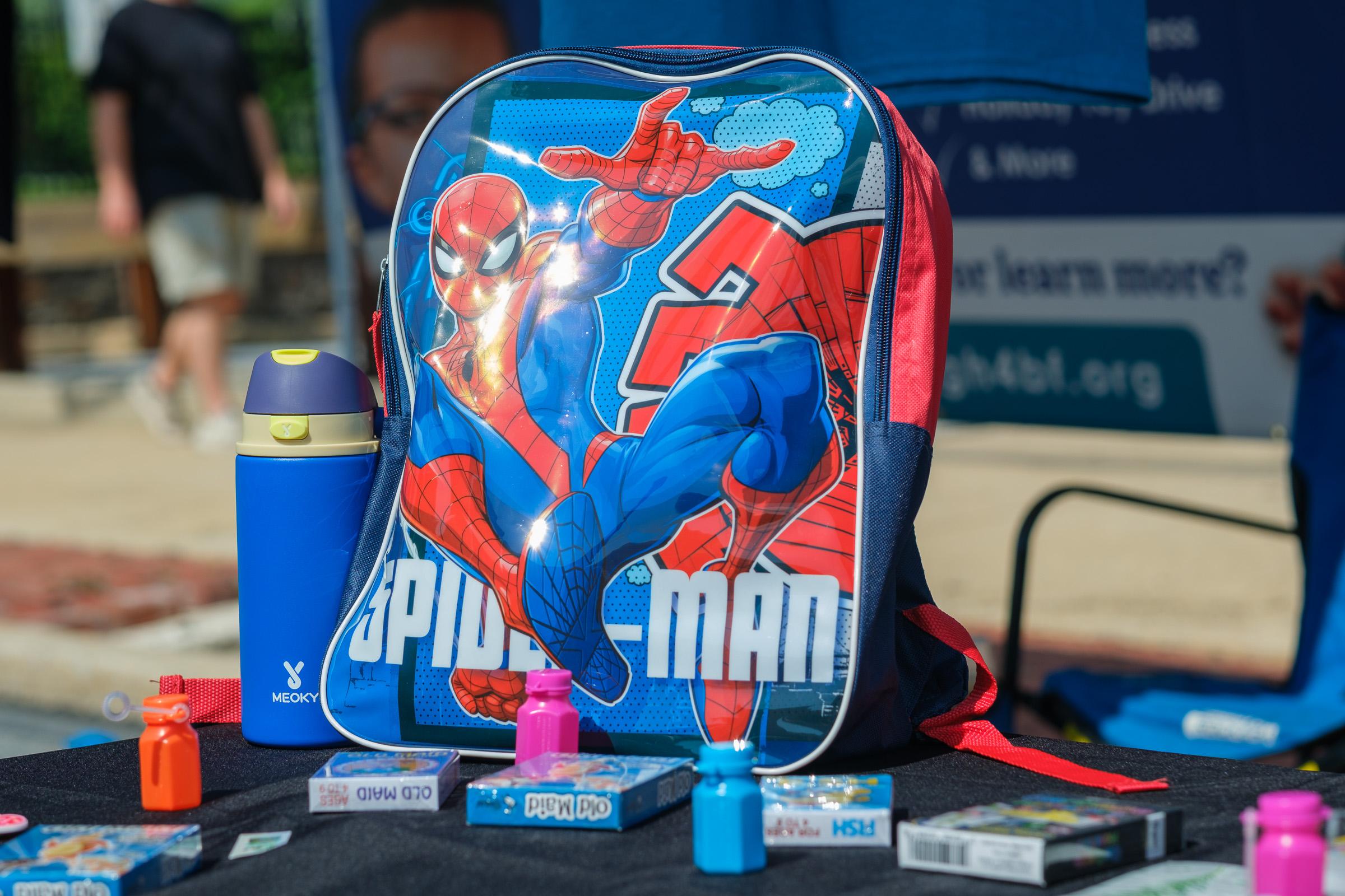 The Stuff-A-Bus giveaway table. A Spider-Man backpack front and centre, a blue Meoky water bottle, bubble bottles in orange, pink and blue, and Old Maid and Go Fish card games spread on a black cloth.