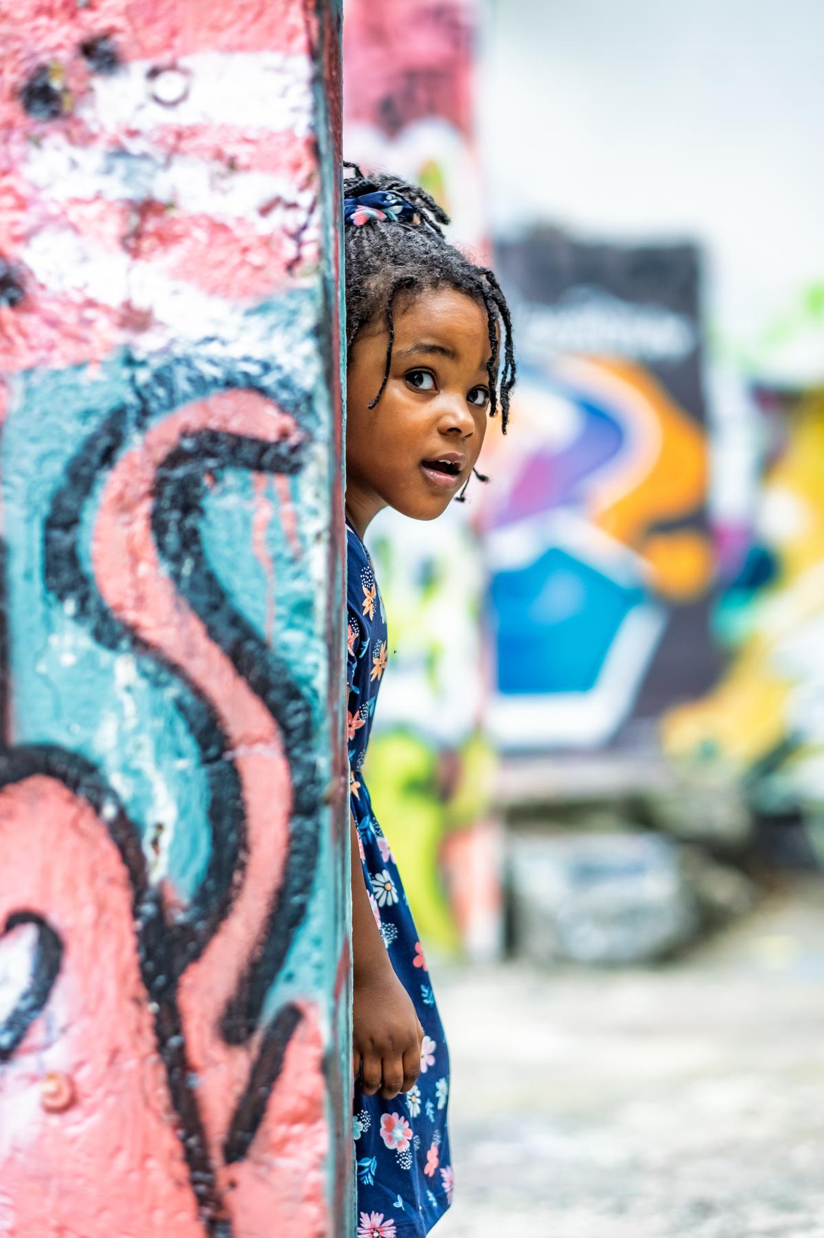 Charlie peeking around a pink and teal painted column, wide-eyed, murals blurred behind her.