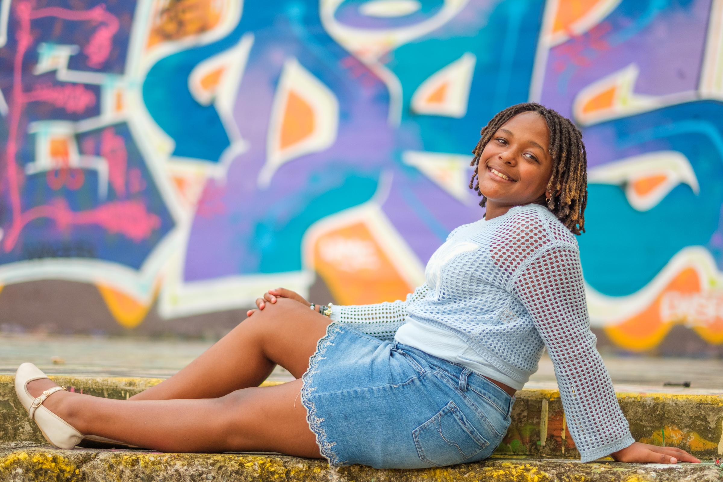 Cori sitting on a low concrete step leaning back on her hands, a blue and purple mural filling the wall behind her.