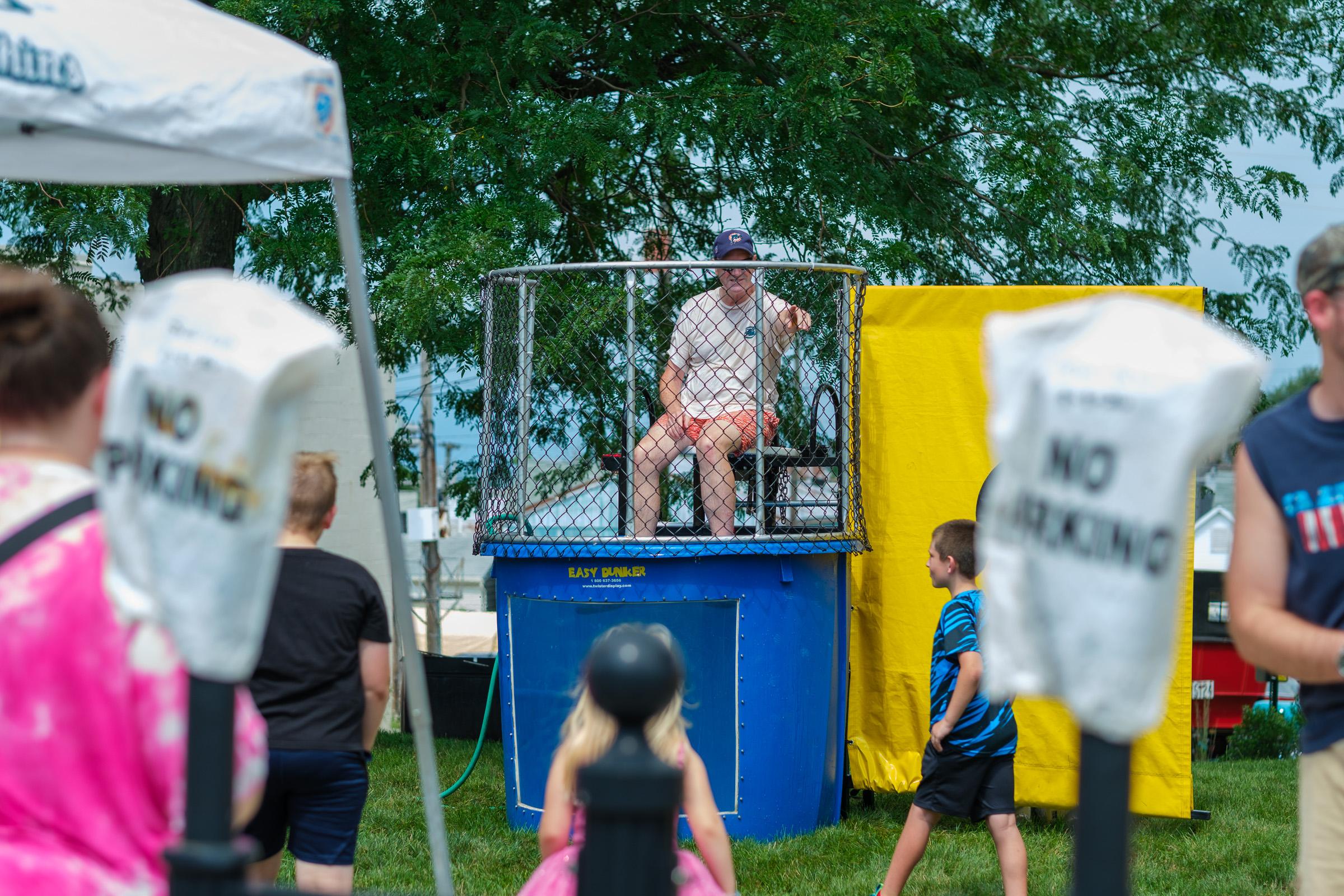 Dunk tank. A man in a ball cap in the cage, kids watching from the grass.