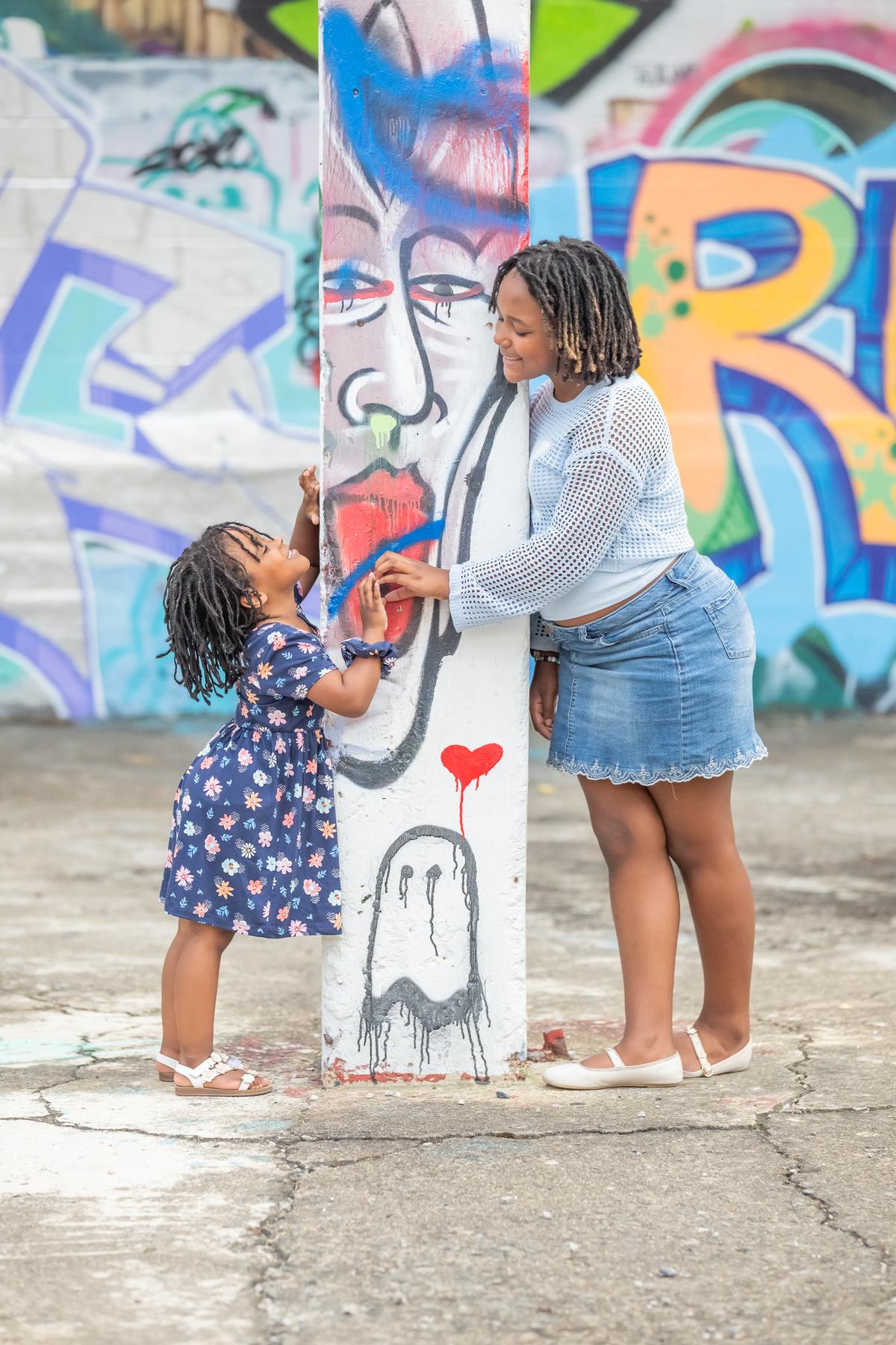 Charlie on tiptoe reaching up a painted column toward Cori, a face mural and a red dripping heart on the column between them.