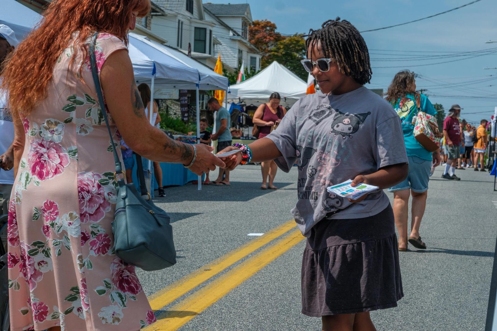 A young girl in sunglasses hands a printed flyer into the outstretched hand of a woman on a closed street lined with vendor tents. She holds a stack of more flyers in her other hand.