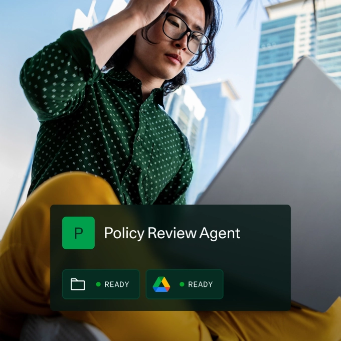 A person wearing a green patterned shirt and yellow pants is working on a laptop outdoors in an urban environment with tall glass buildings in the background. Overlaid on the image is a digital interface titled "Policy Review Agent" with icons indicating folder and Google Drive access, both marked as "READY."