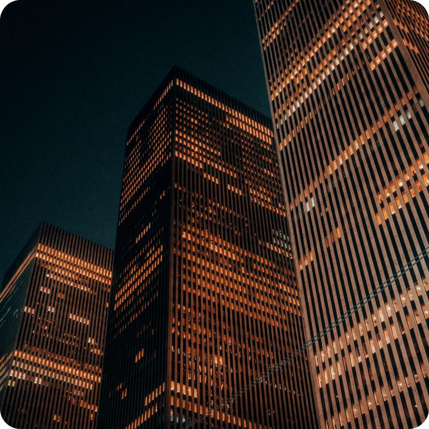 Nighttime view of tall office skyscrapers with illuminated windows, creating a warm orange glow against the dark sky. 