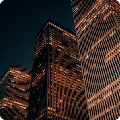 Nighttime view of tall office skyscrapers with illuminated windows, creating a warm orange glow against the dark sky. 
