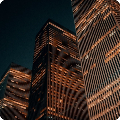 Nighttime view of tall office skyscrapers with illuminated windows, creating a warm orange glow against the dark sky. 