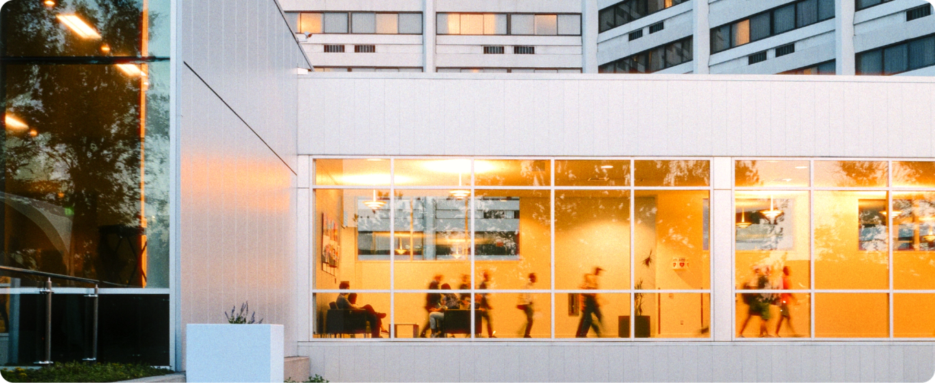 Vue de l’extérieur d’un bâtiment moderne avec de grandes fenêtres qui brillent de mille feux. À l’intérieur, des silhouettes floues marchent et s’assoient, dégageant une atmosphère animée et vivante.
