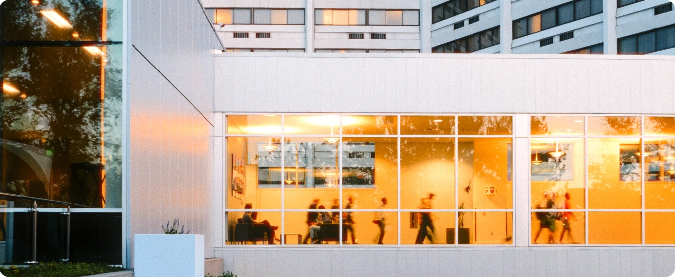View of a modern building exterior with large windows glowing warmly. Inside, blurred figures walk and sit, exuding a bustling, lively atmosphere.