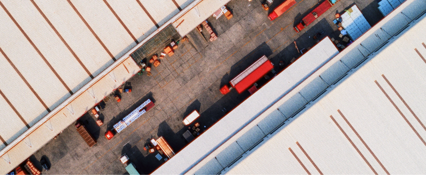 Vue aérienne d’un entrepôt au toit ondulé, entouré de camions rouges et de conteneurs d’expédition. La scène est ordonnée et industrielle.