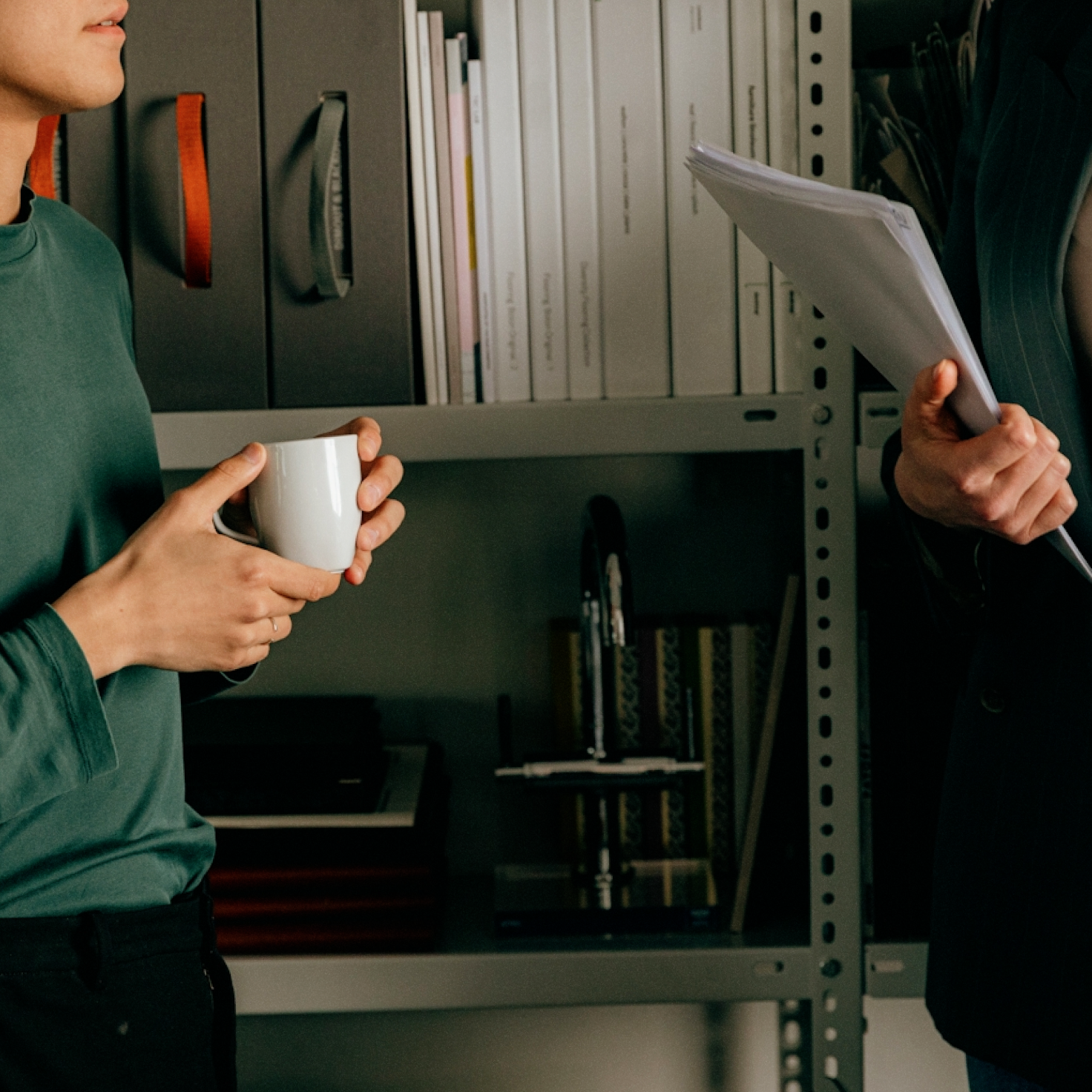 Deux personnes debout devant des étagères de bureau ; l'une tient une tasse blanche, l'autre une pile de documents.