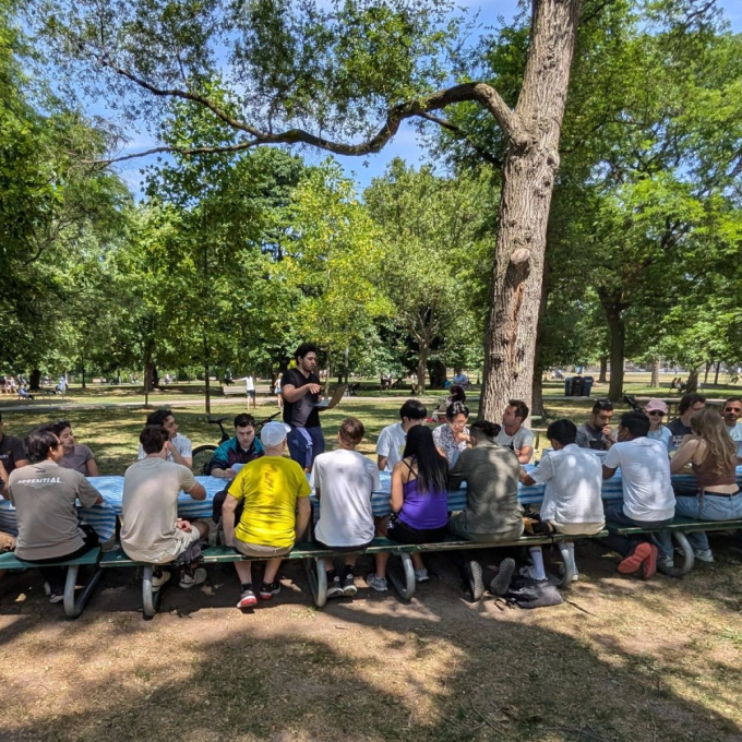 Large group of people sitting together at a long picnic table in a park on a sunny day, listening to a person standing and speaking. Tall trees and green foliage surround the gathering.