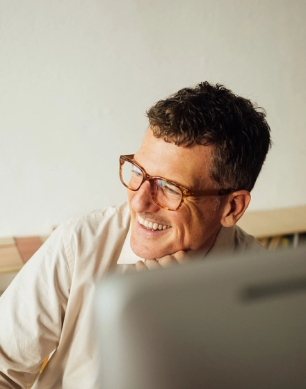 Man wearing glasses and a light-colored shirt smiling while looking off to the side, with part of a computer screen visible in the foreground.