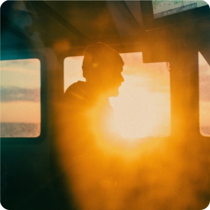 Silhouette of a person wearing a beanie inside a vehicle or cabin, illuminated by the warm glow of a bright sunset shining through the window.