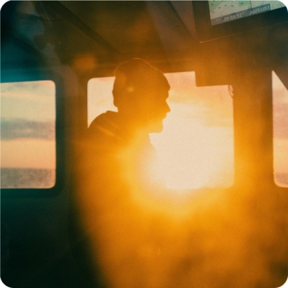 Silhouette of a person wearing a beanie inside a vehicle or cabin, illuminated by the warm glow of a bright sunset shining through the window.