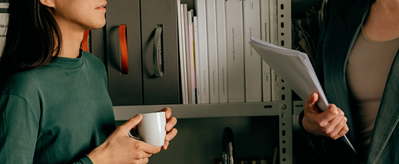 Two people standing in front of office shelves; one holds a white mug, the other holds a stack of documents.