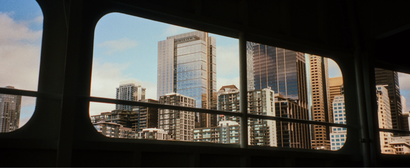 Vue d’un paysage urbain à travers la fenêtre d’un ferry, montrant de grands gratte-ciel modernes sur fond de ciel bleu. La scène est dynamique et urbaine.