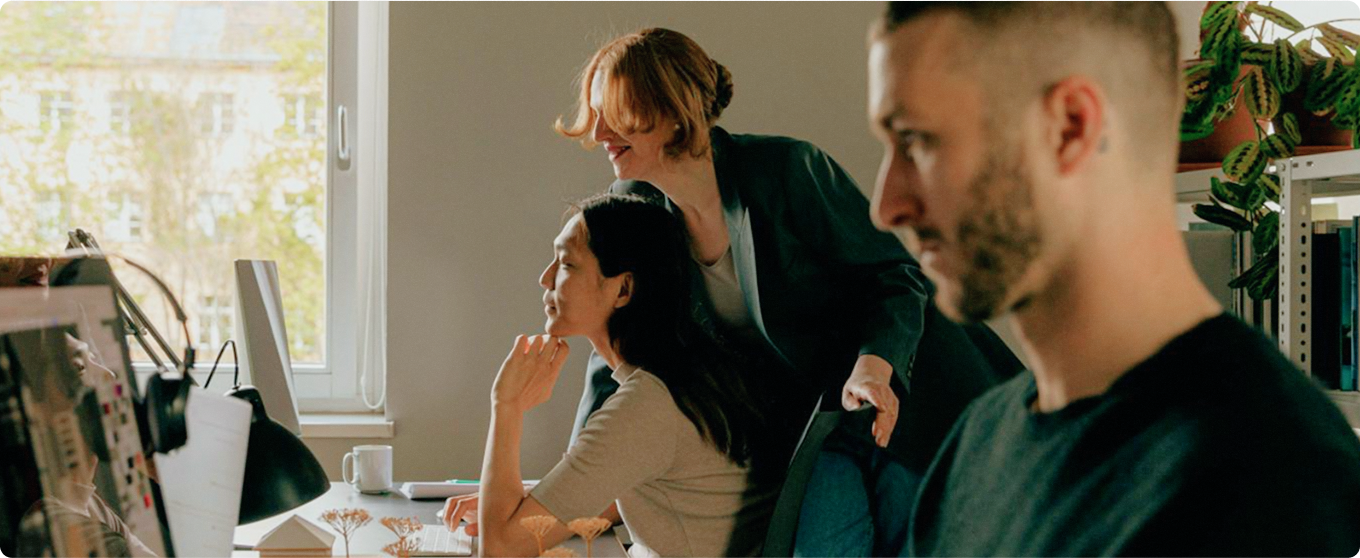 Three people working in an office, two collaborating at a computer while another focuses on his own work in the foreground.