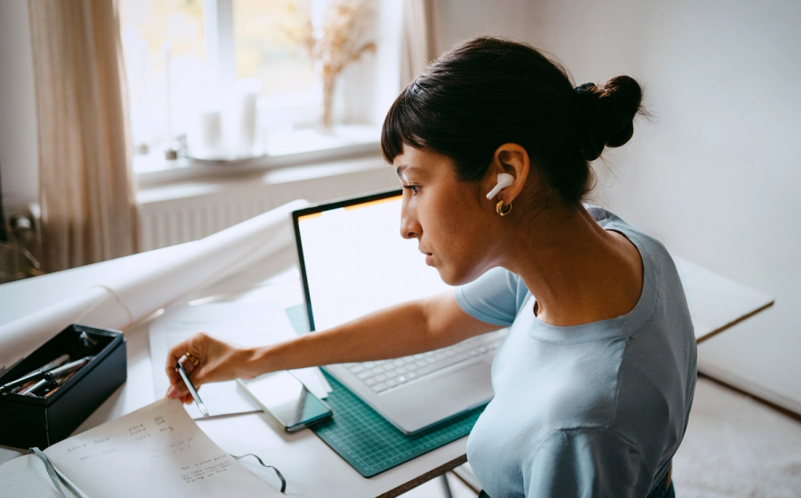 Person wearing wireless earbuds works at a desk with a laptop, writing notes in a notebook while referencing documents.