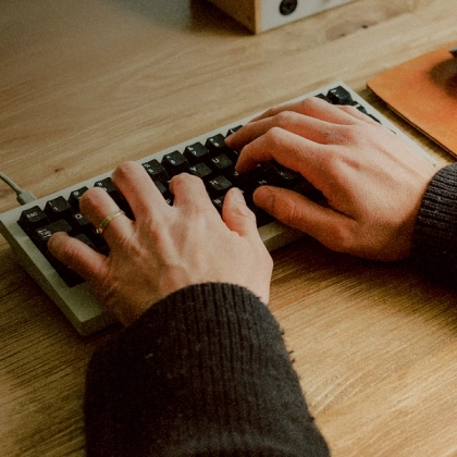 Close-up of a person’s hands typing on a mechanical keyboard on a wooden desk, with part of a notebook and computer equipment visible in the background. 