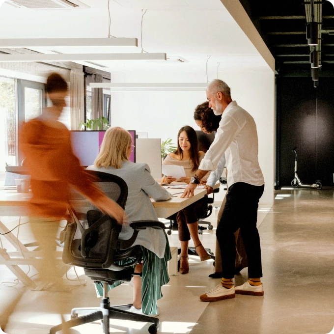 Group of colleagues collaborating at a desk in a modern office, with one person walking past in motion blur.