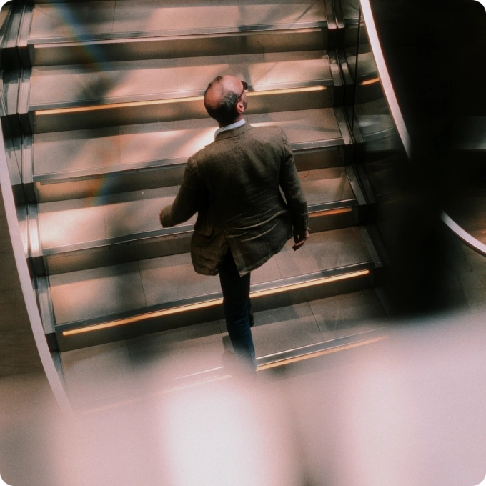 Man in a blazer walking up a modern staircase, viewed from above with soft light reflections.
