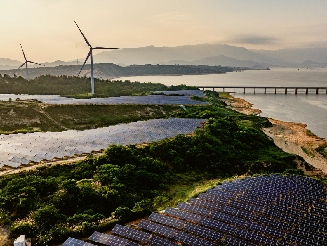 A coastal renewable energy site featuring large fields of solar panels, wind turbines in the background, and a long bridge spanning across the water, set against a backdrop of mountains at sunset.