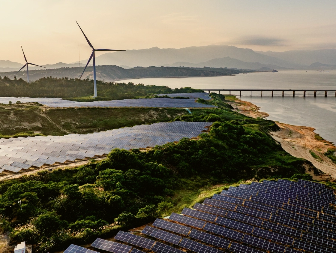 A coastal renewable energy site featuring large fields of solar panels, wind turbines in the background, and a long bridge spanning across the water, set against a backdrop of mountains at sunset.